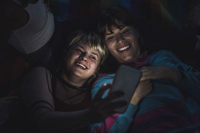 Happy non-binary person taking selfie with friend through mobile phone at night