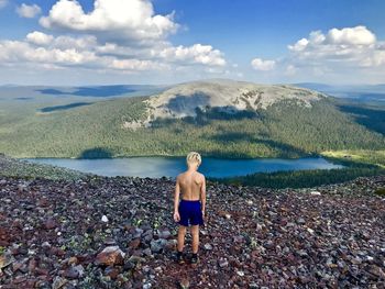 Rear view of shirtless boy looking at lake and mountains against sky