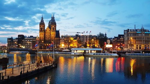 Illuminated buildings by river against cloudy sky