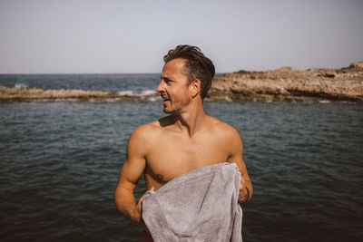 Man looking away while holding towel at beach against sky