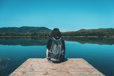 Rear view of man sitting on lake against clear blue sky