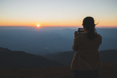 The back view of asian female traveler taking photos of the sunset with a mobile phone