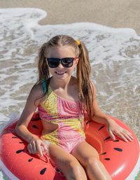 A young girl in sunglasses sits on the sand with an inflatable ring on the shores 