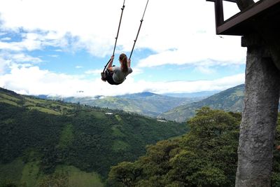 Rear view of young woman swinging above green mountains