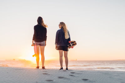 Rear view of women standing with skateboards at beach against sky