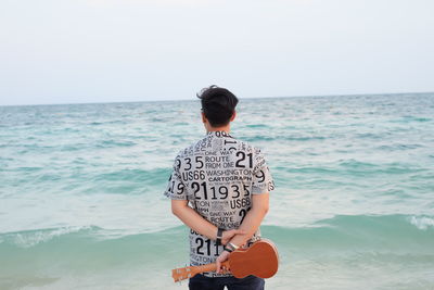 Rear view of young man holding violin while standing by sea against sky