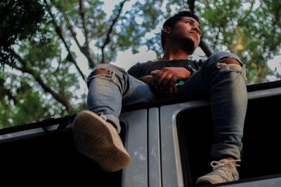 Low angle view of thoughtful young man looking away while sitting on car roof against trees