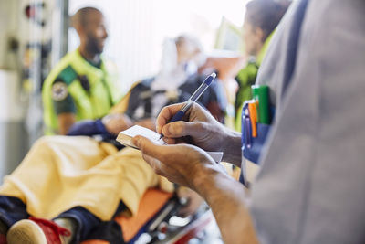 Close-up midsection of male nurse writing medical report against paramedics and patient