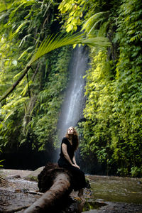 Woman sitting on log against waterfall in forest