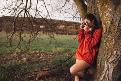 Young woman photographing from camera while standing by tree on land