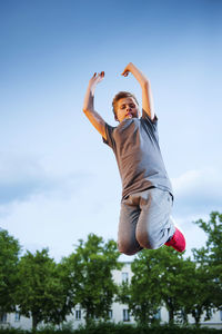 Male teenager jumping in the air with his arms stretched up on the sports field against a blue sky.