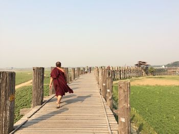 Rear view full length of monk walking on boardwalk against clear sky