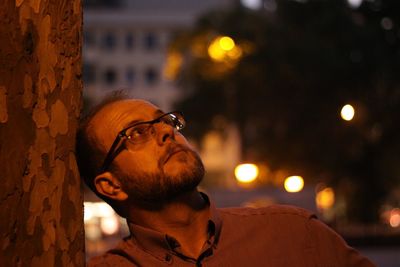 Close-up of mature man looking away while standing by tree trunk at night