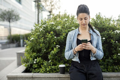 Young woman sitting on retaining wall while using phone in city