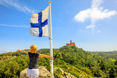 Rear view of woman with arms outstretched standing by flag on mountain against sky during sunny day