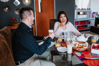 Cheerful young latin american married couple in casual clothes smiling and looking at each other while having breakfast together at home