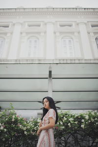 Portrait of young woman standing by plants against building