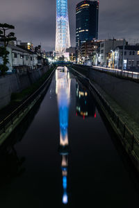 Illuminated bridge over river by buildings against sky at night