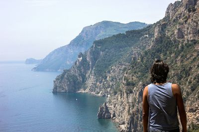 Rear view of man looking at sea by mountains against sky