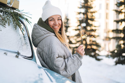 Young happy woman in warm clothes with sparkler in hands near decorated retro car in winter street