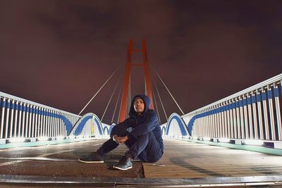 Full length portrait of man sitting on bridge against sky at night