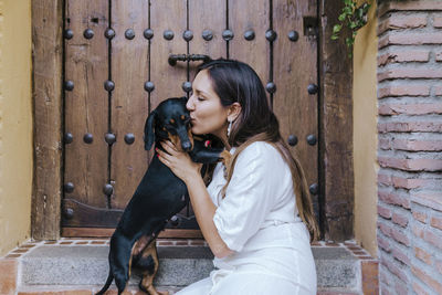 Side view of woman kissing dog while sitting at house doorway