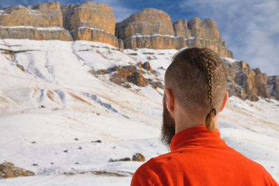 A bearded man with a stylish haircut in a red sweater stands with his back and looks into mountains