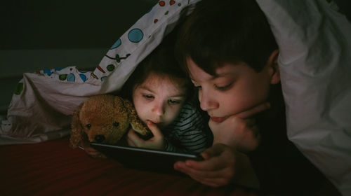 Siblings using digital tablet on bed in darkroom