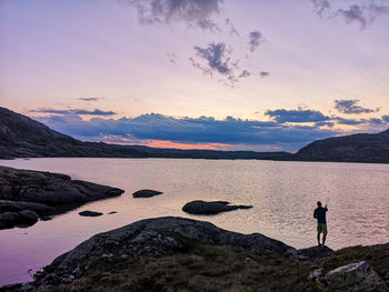 Rear view of silhouette man standing on cliff by lake at sunset