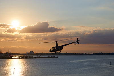 Helicopter flying over river against sky during sunset