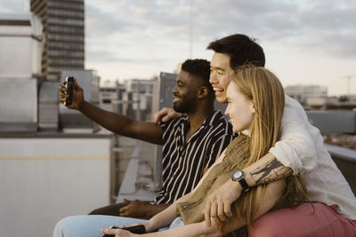 Young man taking selfie with friends through smart phone while sitting on terrace