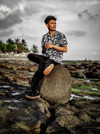 Full length of man sitting on rock looking away against sky