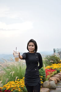 Portrait of smiling young woman holding coffee while standing on grassy field against sky