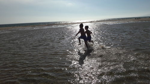 Silhouette siblings running on beach against sky