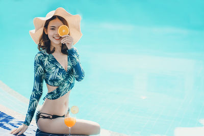 Portrait of woman holding orange while sitting by swimming pool