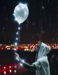 Side view of man holding illuminated helium balloon during snowfall at night