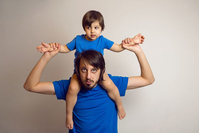 Boy sits on his father's neck in blue t-shirts against the white wall of the house