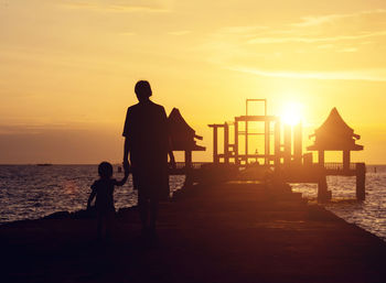 Silhouette boy standing by sea against sky during sunset