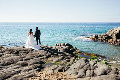 Rear view of newlywed couple standing at rocky beach against clear sky