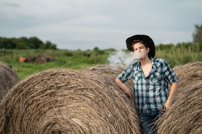 Portrait of a handsome young man in a cowboy hat vaping an electronic cigarette near the haystack.