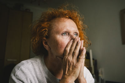 Thoughtful sad senior woman with hands covering mouth sitting at home