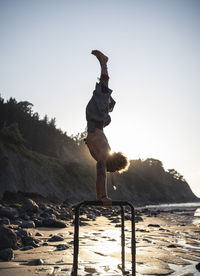 Young man practicing handstand on parallel bars at beach against sky
