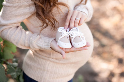 Pregnant woman holding baby boots wearing beige sweater posing outdoors.