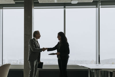 Businessman and businesswoman shaking hands while standing by window at office