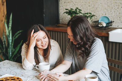 Adult smiling brunette women friends having breakfast and using mobile phone in kitchen at the home