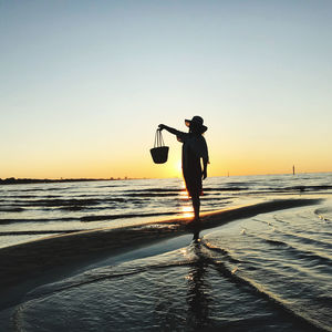 Silhouette woman holding basket standing on beach against sky during sunset