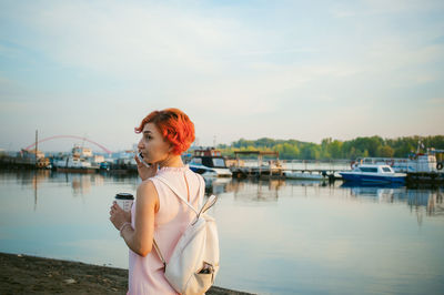 Mid adult woman talking on mobile phone while standing by river against sky during sunset