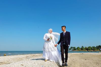 Couple holding umbrella while standing on shore against blue sky