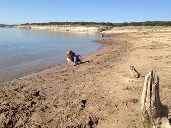 Girl crouching at beach against clear blue sky during sunny day