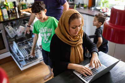 High angle view of woman using laptop on dining table with family working in background at kitchen